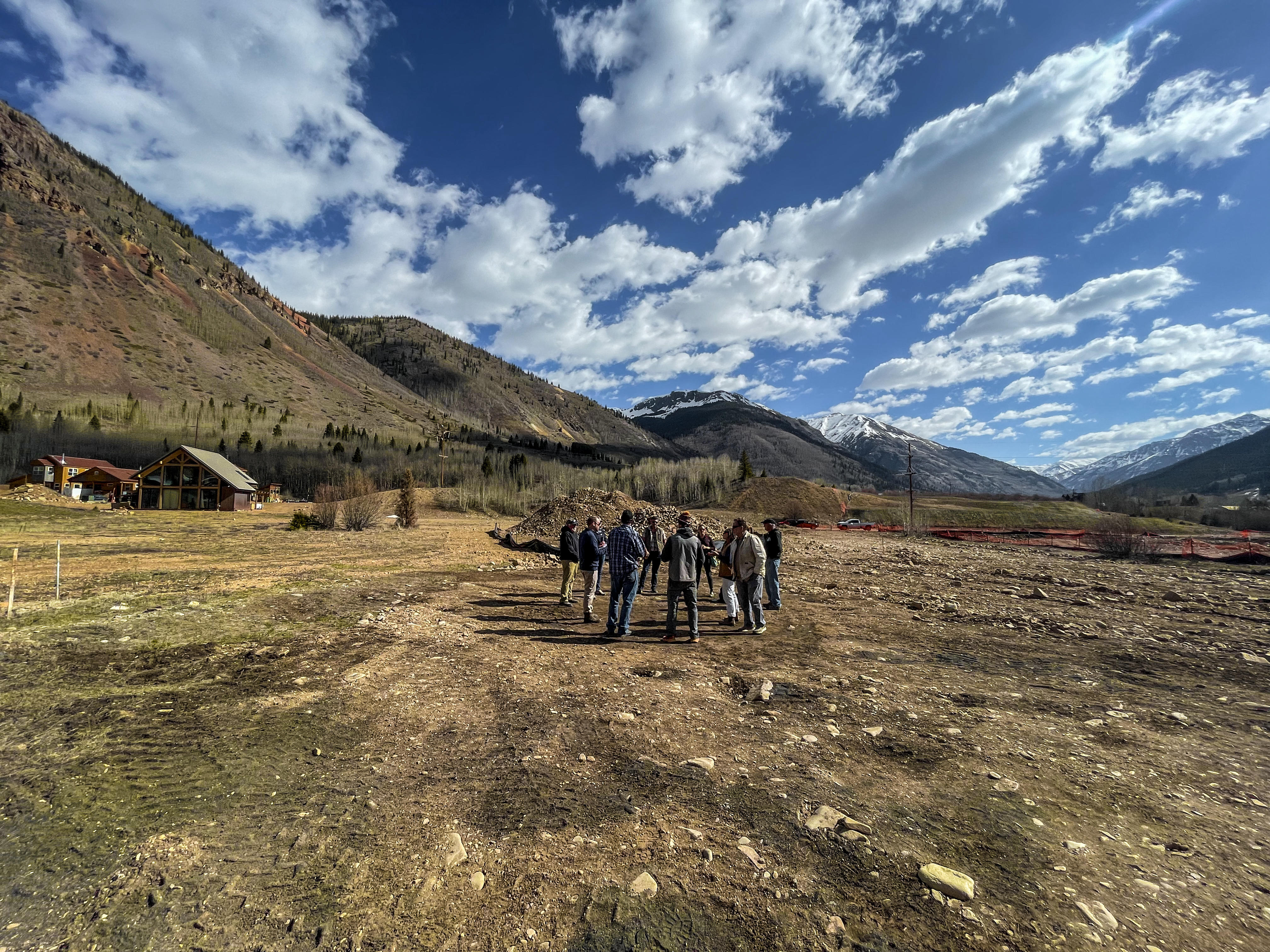 Group of people standing in a circle on a dirt lot with mountains in the background. 